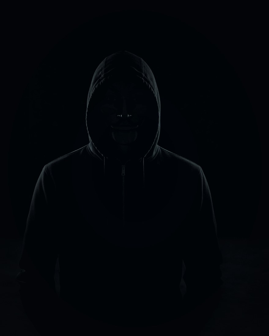 Hooded operator working at a glowing multi-monitor setup, face hidden in shadow